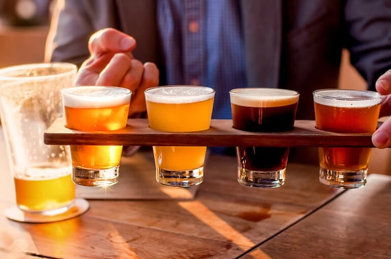 A person holding a wooden flight board with four different craft beers, ranging in color from pale to dark, on a wooden table at a bar with a half-full pint glass in the background.