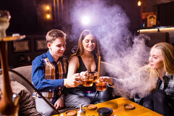Group of friends enjoying beer and hookah together in a lounge, surrounded by warm lighting and smoke
