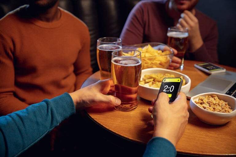 A group of people at a bar enjoying beer and snacks while using a smartphone with a gaming or betting app on the screen.