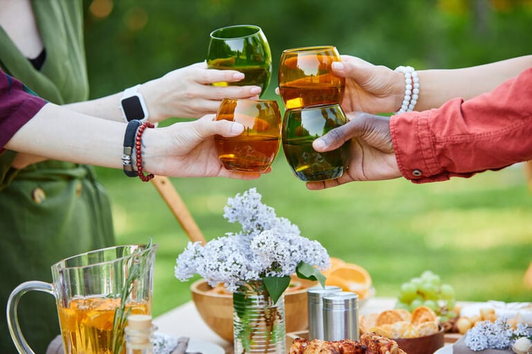 Friends clinking colorful beer glasses over a backyard picnic table with snacks and flowers, showcasing creative ways to serve beer at an outdoor gathering.
