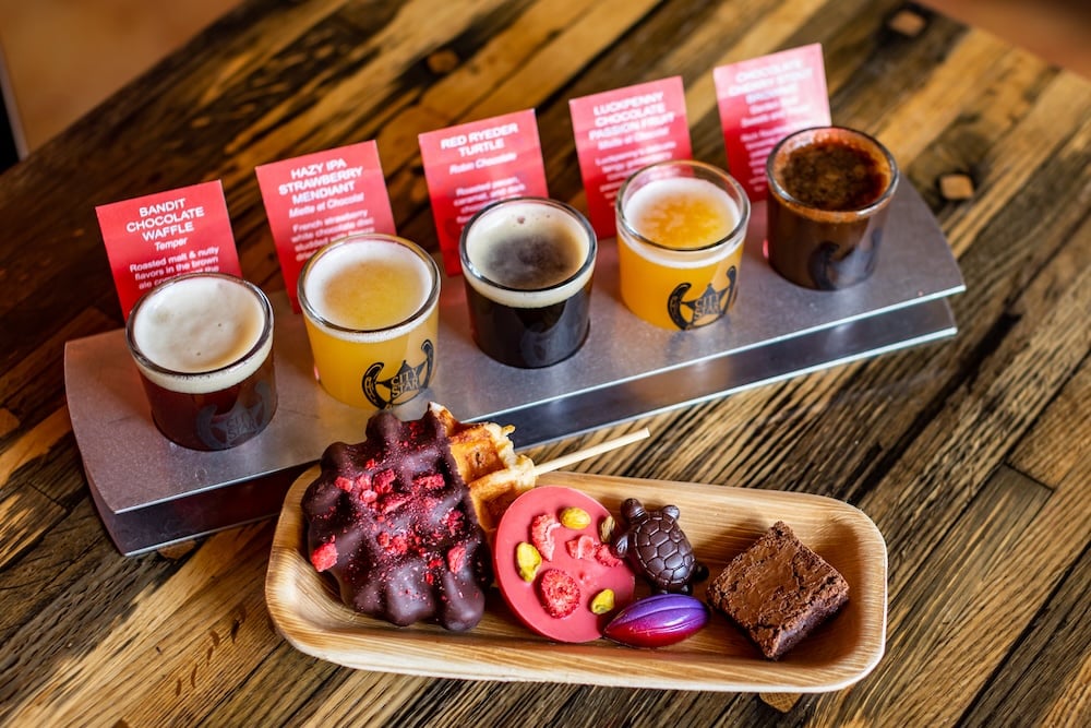 A flight of beers on a wooden table with chocolate pairings and descriptive notes